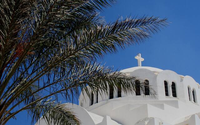 Wit kerkje met koepel en een palmboom tegen een blauwe lucht op Santorini